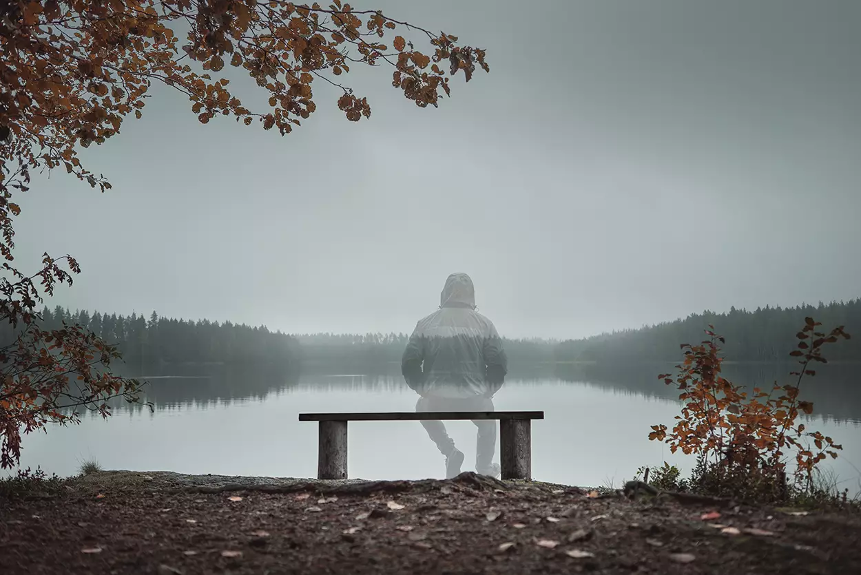 Photo of a man sitting on a bench looing out across a large lake surrounded by trees. The man is faded to represent someone who is no longer there.